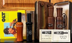 Vintage p.s. olt duck and goose call devices displayed with their original boxes and instruction cards on a wooden shelf.
