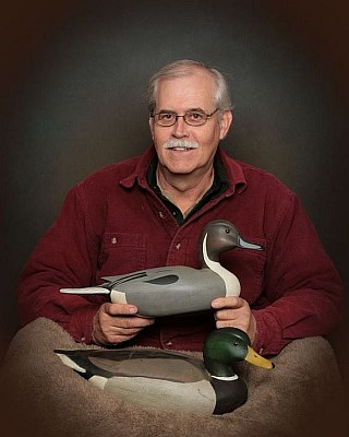 An older man with glasses and a mustache, smiling while holding two wooden duck decoys, one on his lap and another by his side.