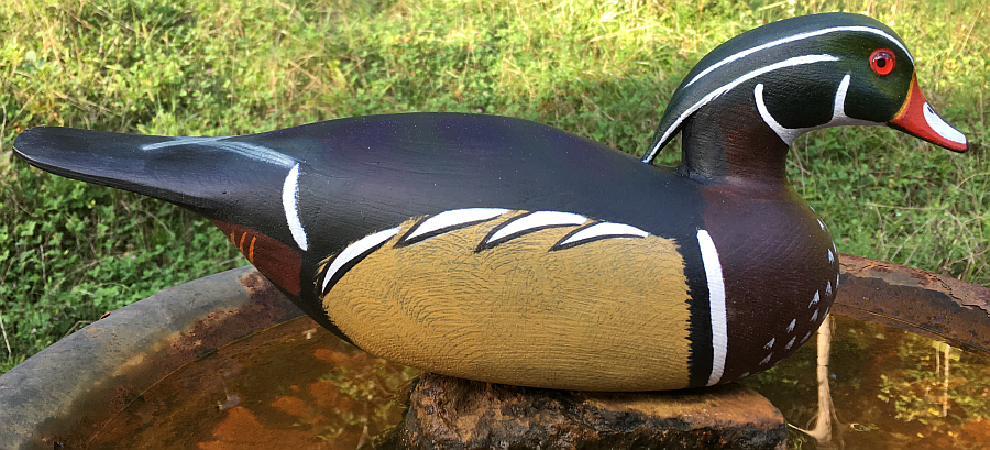 A wooden decoy of a male wood duck, painted with detailed and colorful markings, resting on a rustic metal surface.
